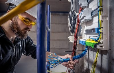 A male electrician works in a switchboard with an electrical connecting cable, connects the equipment with tools.
